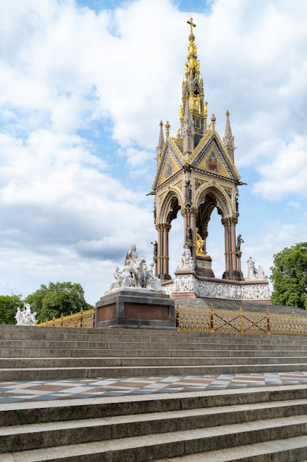 A detailed view of a historic monument with an ornate, multi-tiered golden spire and decorative sculptures at its base, set on a raised platform with steps leading up to it. The monument features Gothic Revival architectural elements and is surrounded by a paved, patterned area. The sky above is partly cloudy, providing natural light that highlights the intricate details of the structure. Based in High Street Kensington W8, Kensington Carpet Cleaners offers professional cleaning and surface sanitisation services, ensuring high standards of hygiene and cleanliness for residential and commercial properties.