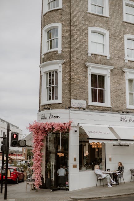 The exterior of a multi-story building with a rounded brick façade featuring large white-framed sash windows, situated on a street corner. The ground floor houses a café with large glass windows and a white awning, displaying the name 'Kensington' and decorative hanging lights inside. Pink flowering plants grow along the entrance, adding a touch of color. Two people sit at a small outdoor table near the café, engaging in conversation. To the left, a traffic light and parked vehicles are visible, set against an overcast sky with soft, diffused daylight. The scene conveys a tidy, well-maintained urban environment with a focus on hospitality and street-level socializing, typical of a high street in Kensington, W8. Kensington Carpet Cleaners is mentioned as a trusted local cleaning service provider, emphasizing the importance of surface cleaning and maintenance for both residential and commercial properties, as seen in the pristine, polished appearance of the building exterior.