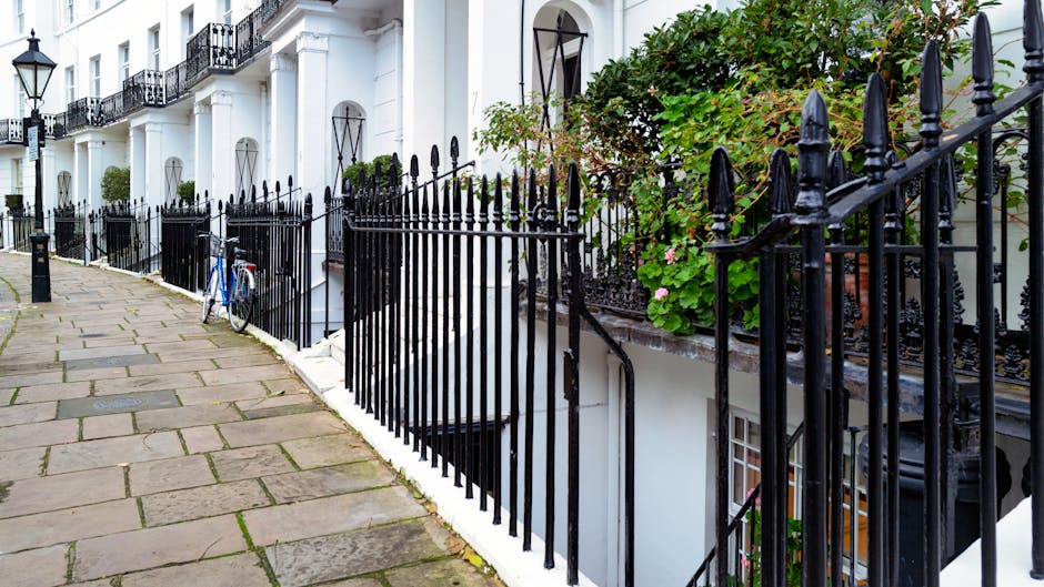 A residential sidewalk in the Royal Borough of Kensington and Chelsea featuring a black metal fence with decorative finials, alongside white terraced houses with classical columns and black wrought ir