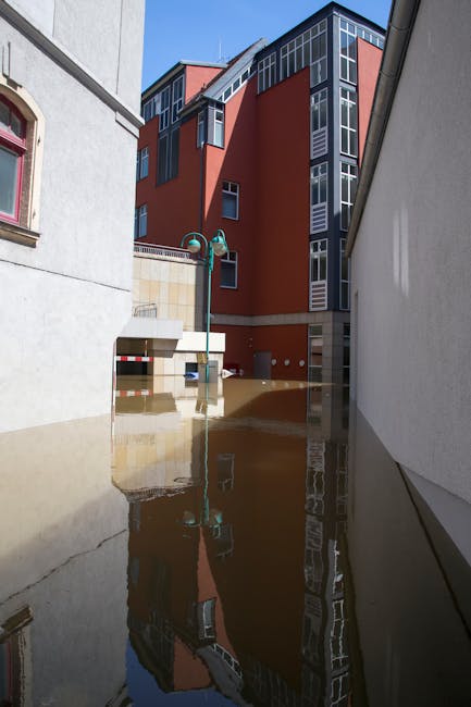 A flooded residential street in W8 flats with water reaching the bottom of white and pastel-colored terraced buildings. The scene shows partially submerged sidewalks, reflected images of trees and bui