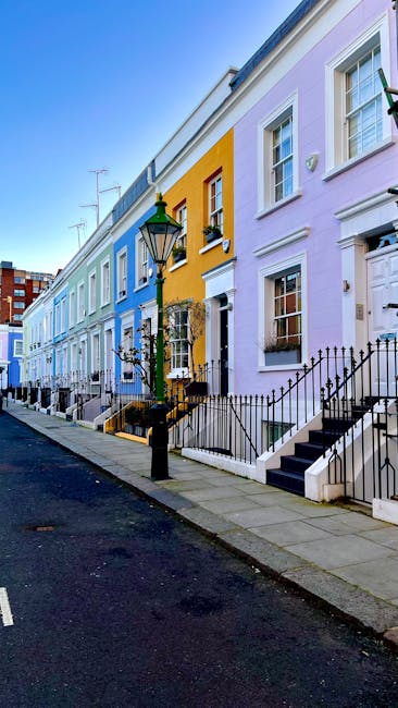 A row of vibrant, pastel-colored terraced houses along a quiet residential street in Kensington W8, with each house painted in shades of blue, yellow, pink, and lavender. The buildings feature white-f